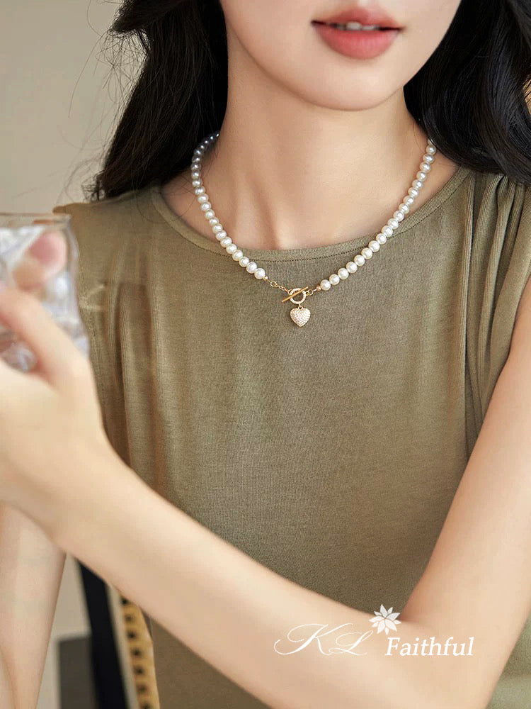 Woman wearing a pearl necklace with a heart pendant, holding a glass of water.